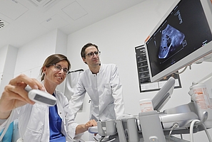 Es sind zwei Mediziner im weißen Kittel zu sehen. Die Ärztin hält ein Ultraschallgerät in der Hand, sie und ihr Kollege schauen auf einen Monitor. 
